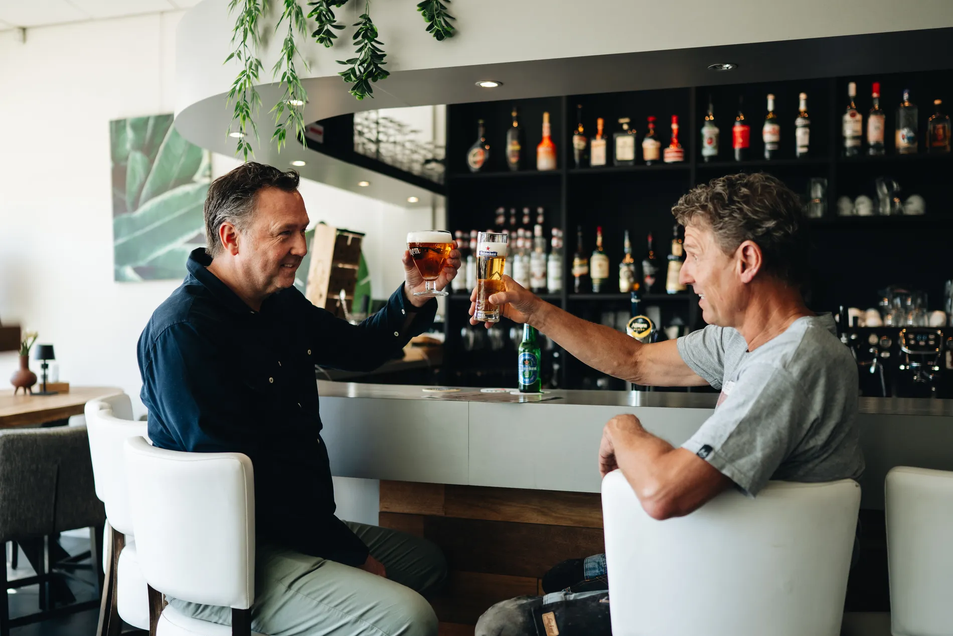 Twee mannen die aan de bar van het restaurant zitten en glimlachend met elkaar proosten met een glas getapt bier.